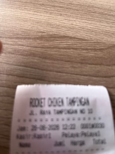 The image shows a receipt from "ROCKET CHIKEN TAMPINGAN" which is located at "JL. RRAYA TAMPINGAN NO 10". The receipt contains a transaction date of "26-06-225 12:22" and a transaction number "00010000". The receipt also has columns for "Jual Harga" and "Total", but the specific items and prices are not legible. The receipt is placed on a wooden surface, possibly a table. There are no people visible in the image.