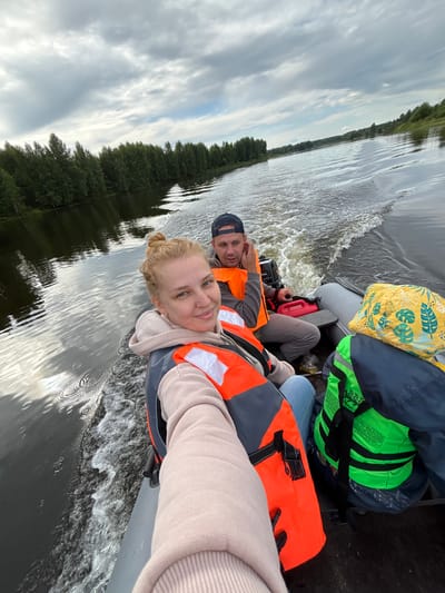 A selfie is taken from an inflatable boat on a body of water, likely a river, with a man and a woman aboard. The woman, in the foreground, smiles at the camera, her blonde hair pulled up in a bun. She wears a light pink hoodie and an orange life vest over it. The man, behind her, is wearing a dark baseball cap and an orange life vest. He is looking towards the right, his hand near his ear. In the background, a forested shoreline lines the river. The water is choppy, indicating the boat is moving. Luggage, including a yellow patterned bag and a bright green life vest, is visible in the boat. The sky is overcast with dramatic clouds. The scene is set in Anteplevo (Okhotinskoe S/P), Russia.