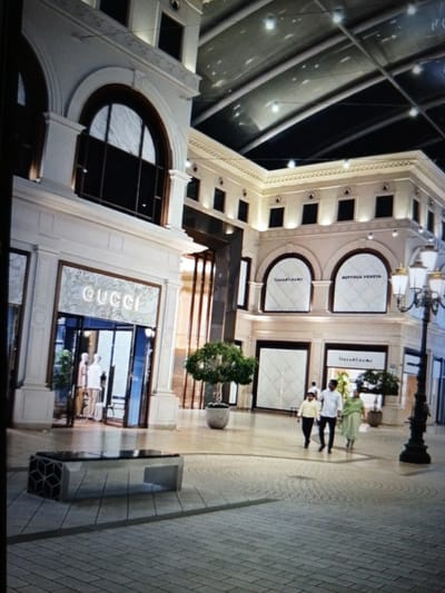 The image depicts the interior of a high-end shopping mall, possibly in Golbembal, Nigeria. The architecture is grand, with large arched windows and a ceiling with visible structural beams and lighting. White and cream colored facades dominate the scene, with ornate detailing along the upper levels.
The primary subjects are three people walking across the tiled floor of the mall. They appear to be a family unit, with two adults and one child. The adult male is wearing a white shirt and dark pants, the adult female is in a light green outfit, and the child is in a light yellow shirt and dark pants. Their expressions are not discernible due to the image quality and distance, but they appear to be casually walking through the mall.
Visible text includes "GUCCI" displayed prominently on the facade of one of the shops. Another shop's signage appears to be in Arabic script, and another displays "BOTTEGA VENETA". This suggests a luxury retail environment.
Notable details include the polished tiled flooring, a modern bench with a geometric pattern at its base, and potted plants decorating the space. The lighting suggests it is daytime, or at least well-lit indoors, though no natural light or indication of weather is apparent. The overall atmosphere is one of upscale shopping and leisurely movement within a well-maintained interior space.