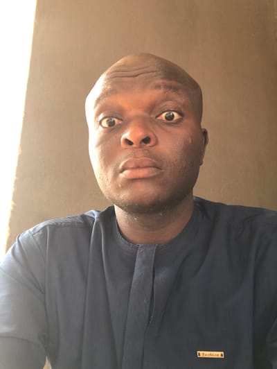 An adult male with dark skin and a bald head is depicted in a close-up, head-and-shoulders portrait against a plain, light brown background. He is looking directly into the camera with wide, open eyes and slightly parted lips, exhibiting visible forehead wrinkles. He is wearing a dark blue traditional garment with long sleeves and a round neckline featuring a short placket. A small, rectangular, gold-colored label inscribed with "Fashion" is affixed to the garment's left chest area. A strong light source illuminates the scene from the subject's left, creating a bright area on the upper left of the background and highlighting the left side of the subject's face. The scene is located in Egboje, Nigeria.