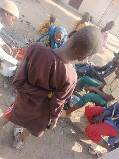 An outdoor scene in Gobirawa, Nigeria, depicts approximately nine individuals gathered on a dusty, unpaved ground surface. The foreground is dominated by the back of a light-skinned individual wearing a long-sleeved, brown tunic, a gold-colored bracelet on the right wrist, and a single white Adidas slide on the left foot. This person's head is cropped above the eyes, showing short, dark hair.

To the left, another individual in a white garment and light-colored cap attends to a white plastic container, near which a red plastic container and a bundle of sticks are visible. Further left and center, an elderly woman with dark skin, wearing a blue headscarf and a green patterned garment, crouches, looking towards the left with a faint smile.

In the mid-ground and background, several other individuals are present. One person in a yellow garment and cap crouches, holding a blue plastic item. Another person in a yellow garment stands partially obscured. Two individuals sit on the ground to the right: one in a light-colored shirt with green details and green pants, with a silver metal bowl nearby; the other in a blue garment over red pants. Both are wearing light-colored slides. A black plastic bag is positioned between them.

The environment includes distant light-colored building structures with minimal architectural detail. The ground is dry earth, suggesting an arid climate. Strong shadows indicate bright daylight conditions. There are no clear actions of direct interaction between all individuals, but they are positioned in close proximity, suggesting a communal gathering.