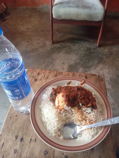 The scene is depicted indoors in Okere-Urhobo, Nigeria, focusing on a meal presented on a wooden surface. In the foreground, a round, light-colored plate with a brown rim contains white rice topped with a halved hard-boiled egg and a reddish-orange stew with visible green elements. A silver-colored metal spoon rests within the plate. Adjacent to the plate, a clear plastic bottle of Aqua Rapha brand drinking water stands upright. The supporting surface is a wooden table composed of rough planks, exhibiting prominent grain and wear. In the background, a chair with a reddish-brown wooden frame and a worn, light beige upholstered seat is partially visible. The floor is a light brown concrete or tiled surface with darker discolored areas. A plain, reddish-orange wall is present in the upper left background. No people are visible, and no actions or interactions are occurring.