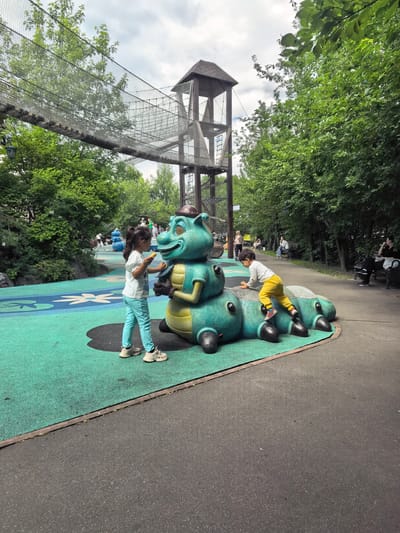 The image depicts a playground in Sovkhoza imeni Lenina, Russia. In the foreground, two children interact with a large, teal-colored caterpillar-shaped play structure. A young girl on the left, wearing a white t-shirt and teal pants, holds a yellow object near the caterpillar's face. To the right, a young boy in a white shirt and yellow pants is climbing on the caterpillar. The playground surface is green with blue and white floral patterns. In the background, a rope bridge connects to a wooden tower structure, surrounded by lush green trees. Several other people are visible in the background, some sitting on benches and others walking. The sky is overcast with clouds.