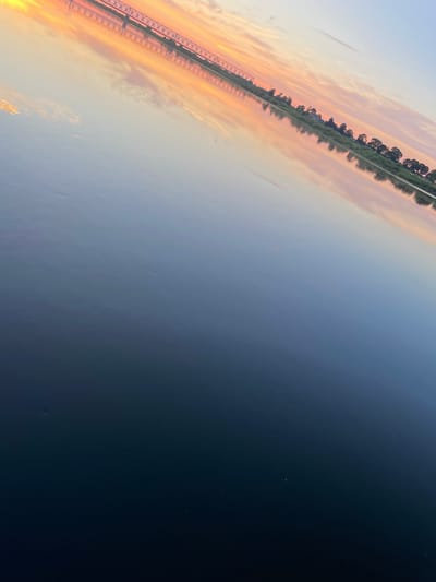 An outdoor shot captures a river at sunset. The sky is awash with hues of pink, orange, and yellow, with scattered clouds adding texture. A long bridge, with intricate metalwork, stretches across the horizon, its reflection mirrored in the calm water. The riverbank is lined with lush green trees and vegetation. The water in the foreground is dark and still, gradually transitioning to a lighter blue as it recedes into the distance. The image is taken at an angle, giving the impression of looking across the water. The scene is located on the river Neman in Sovetsk, Russia.