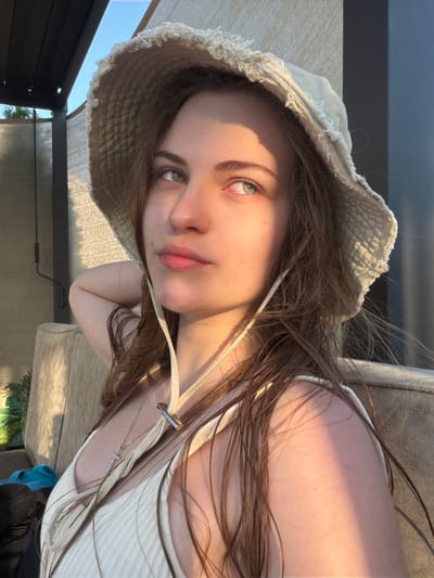 A young person with long brown hair and green eyes, wearing a wide-brimmed tan hat and a white tank top, is lying down outdoors. The sun is casting a warm glow and shadows across their face and body, suggesting it is late afternoon or early evening. They are wearing a silver necklace with a pendant. The setting appears to be a lounge chair or daybed, possibly on a patio or balcony, with a dark structure and some greenery visible in the background. The sky is a clear blue. The overall mood is relaxed and serene. There is no visible text.