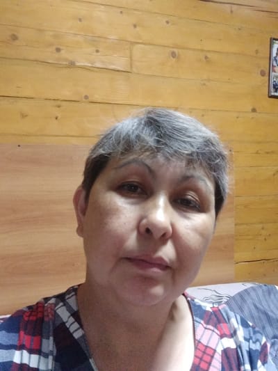A middle-aged woman with short, gray hair is shown from the chest up, looking directly at the camera. She has light-colored skin with some blemishes and is wearing a red, white, and blue plaid shirt. The background is a wooden wall, likely paneling, with horizontal planks of varying shades of yellow and brown. The wood has visible knots and grain. To the right side of the frame, a portion of a framed picture is visible on the wall. The lighting appears to be artificial and the overall scene suggests an indoor setting, possibly a home. The woman's expression is neutral. No text is visible in the image.