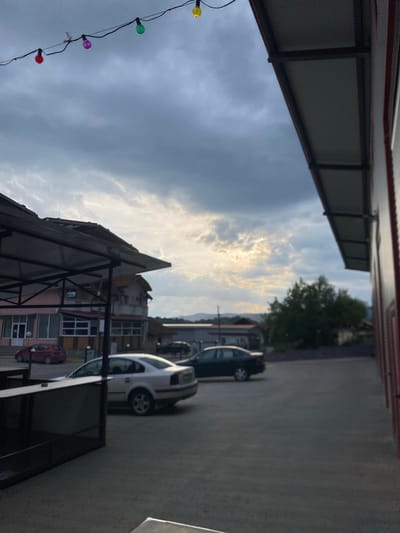 This outdoor image captures a street or parking area in Satovcha, Bulgaria, under a dynamic, partly cloudy sky. The perspective is angled slightly upwards, revealing various urban elements.

The sky dominates the upper and left portions of the frame, showcasing a mix of grey and white clouds. Patches of bright sunlight pierce through the clouds in the upper-middle, illuminating their edges and suggesting either late afternoon/early evening or early morning.

In the upper left foreground, a dark, angled roof structure, possibly an awning or part of a building's roof, is prominent, featuring corrugated material and a supportive dark frame. Below this, a string of colorful decorative lights—yellow, green, purple, and red—hangs against the cloudy backdrop. Another dark, angled structure is visible in the lower foreground, partially obscuring the view of buildings.

To the right, a paved asphalt area serves as a parking space for several vehicles. A white car is visible in the mid-right, while a darker car is parked further back. In the background, a cluster of multi-story buildings with varied facades and colors forms the cityscape, alongside a large, green deciduous tree. A small red car is also parked near one of these buildings. The far right edge of the image is bordered by another building structure.

The scene appears calm and devoid of immediate human activity. No discernible text is clearly visible on any signs or buildings due to the distance and image resolution. The colorful lights introduce a decorative or festive element to an otherwise ordinary streetscape.