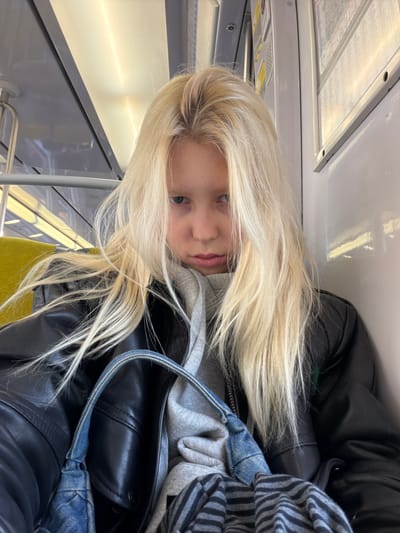 The image displays a single individual, appearing as a young female, seated within what is identifiable as a public transport carriage. The individual has long, straight, platinum blonde hair that falls over her shoulders, fair skin, and light-colored eyes, directed towards the camera with a neutral expression. She is dressed in a grey hooded sweatshirt, over which a black leather-style jacket is worn. A denim shoulder bag is present on her lap, partially covering a black and grey striped fabric object.
The interior environment of the vehicle features a yellow-green upholstered seat to the left of the individual. Overhead, linear fluorescent lighting fixtures illuminate the space. The walls and ceiling are composed of light grey or white panels. A horizontal metal grab bar runs above the seating area. On the wall to the individual's right, a white panel displays what appears to be a map or informational poster, secured by two visible fasteners. The design and elements of the carriage are consistent with those found in RER or Métro trains operating within the Île-de-France region, which serves the city of Arcueil, France. No specific signage or external landmarks are visible to pinpoint the exact location within Arcueil.