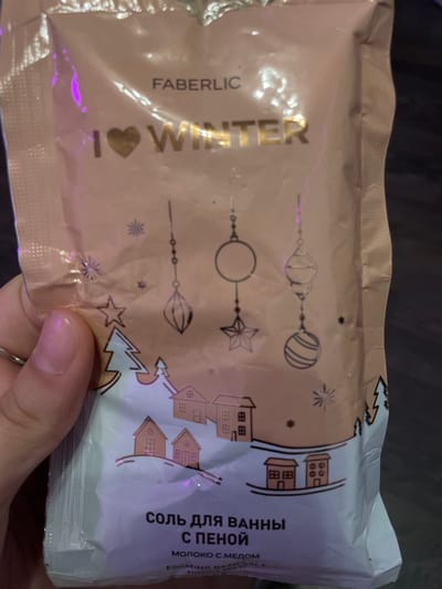 A person is holding a package of bath salt. The package is light brown with white and gold lettering and graphics. The lettering says "FABERLIC" and "I LOVE WINTER". There are drawings of Christmas ornaments, snowflakes, trees, and houses on the package. The bottom of the package has Russian text that translates to "BATH SALT WITH FOAM" and English text that says "FOAMING BATH SALT HONEY MILK". The background is dark and appears to be an indoor setting, possibly a home. There are no discernible landmarks or environmental features that would suggest a specific location within Romanovka, Russia.