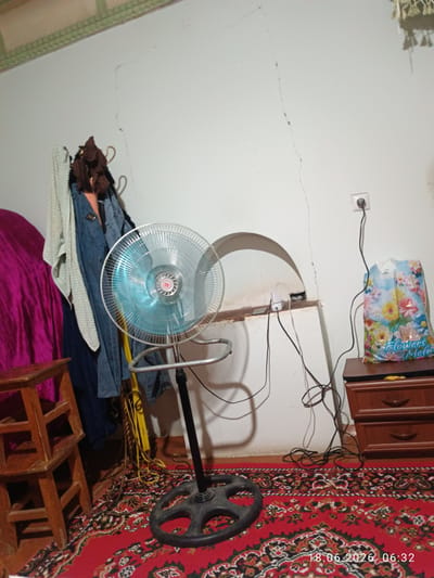 The image depicts an indoor scene, likely a residential room in Zangiata, Uzbekistan. A floor fan with a blue-tinted fan guard is centrally positioned on a black base, with its power cord extending to an outlet on the wall. To the left of the fan, a wooden stepladder with two steps is visible. Behind the fan and stepladder, clothing items, including a denim jacket and a light-colored garment, are hanging on hooks on the wall. A pink fabric is draped on the far left. The floor is covered with a red patterned rug. To the right of the fan, on a wooden dresser with two drawers, sits a plastic shopping bag adorned with floral patterns. The wall exhibits noticeable cracks, and a shelf unit with an arched opening is integrated into the wall above the dresser. An electrical outlet is mounted on the wall near the dresser. The overall impression is that of a cluttered but functional living space.