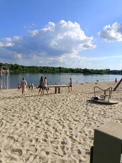 This image depicts a lively summer scene at a sandy beach located by a body of water, likely a lake or river, in Gubkin, Russia. The beach is populated with people enjoying various activities under a bright, partly cloudy sky. In the foreground, children and adults are scattered across the sand, some walking, others sitting or playing. A seesaw made of logs is visible in the middle ground, with two individuals engaged on it. To the left, a lifeguard tower stands tall with a person on top. A merry-go-round, a piece of playground equipment, is situated on the right side of the beach. The water is calm with several individuals swimming or wading. The background features a dense line of green trees, forming the border of the water and creating a natural landscape. The overall atmosphere is one of leisure and recreation on a warm, sunny day.
