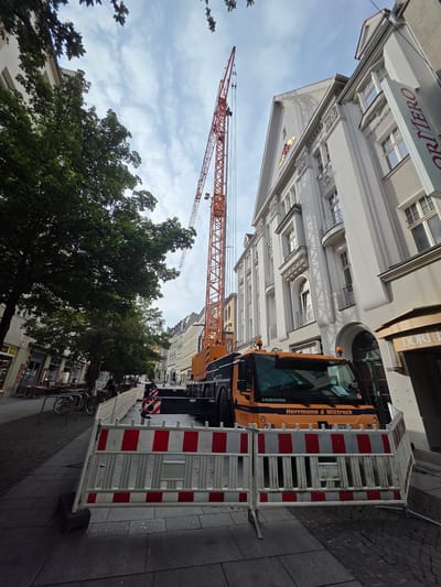 This is a photograph of a street scene in the city of Halle (Saale), Germany. In the foreground, a red and white striped construction barrier is set up on a paved sidewalk. Behind the barrier, a large orange mobile crane is parked on the street, its boom extending upwards into the sky. The crane is identified with the text "Herrmann & Wittrock" and "LIEBHERR" on its cabin. The street is lined with multi-story buildings, some with intricate architectural details and balconies. Trees with lush green foliage are also visible along the sidewalk, casting shadows on the ground. Several bicycles are parked near the barrier. In the background, people are walking along the street, and the facade of a building with a sign that reads "ORTIMERO" is visible. The sky is partially cloudy. The overall scene suggests ongoing construction or maintenance work on the street or in one of the buildings.