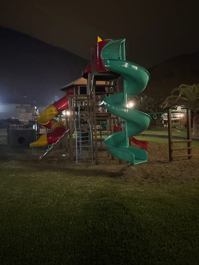 A playground structure with slides, a ladder, and a platform is visible at night in a grassy area. A large green spiral slide is prominent on the right side of the structure. A red and yellow tube slide is on the left. The playground appears to be made of metal and plastic. The background shows some buildings and palm trees, with a dark hill or mountain in the distance. The scene is lit by artificial lights, creating some shadows and highlights. This location is in San Antonio, Peru.