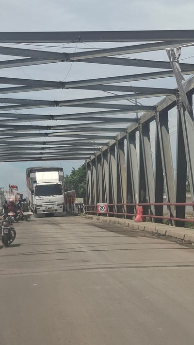 The image depicts a view from a vehicle on a paved road leading onto a metal truss bridge. The bridge structure is prominent, with heavy steel girders forming a geometric pattern overhead and vertical supports along the side of the road. A white truck with a covered cargo area is the most significant vehicle in the frame, positioned on the bridge. To the left of the truck, several motorcycles are visible, with riders on them, suggesting traffic congestion. A red speed limit sign indicating "20" is posted on the right side of the road, near the bridge railing, which is painted red. The environment appears to be a roadside with some vegetation visible beyond the bridge on the right. The sky is overcast with a hazy, pale blue color. The road surface shows signs of wear and tire marks. The perspective suggests the image was taken while moving on the road. The location is identified as the city of Kulukuta, Indonesia.
