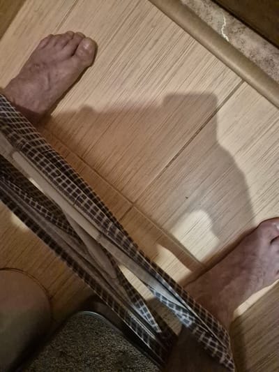 The image shows a pair of men's patterned underwear draped over a person's legs, with their feet visible on a light-colored wood-grain floor. The underwear is primarily black and white plaid. One foot is in the foreground on the right, and another is visible in the upper left corner. A shadow is cast on the floor. The environment appears to be an indoor setting, possibly a bathroom or bedroom, given the tiled floor and the presence of what looks like a toilet seat in the lower left corner. The image does not provide any information to determine the location in Konakovo, Russia.