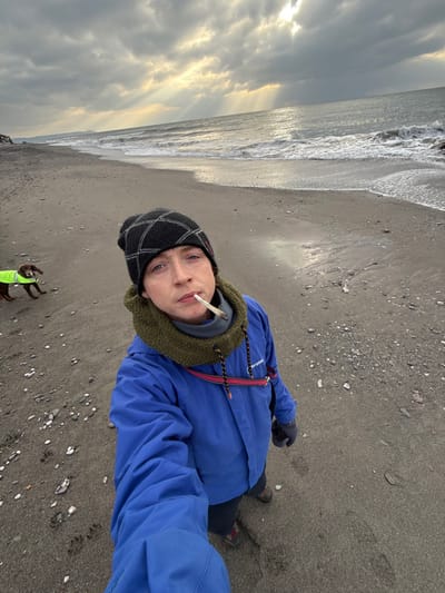 An individual is depicted taking a selfie on a grey-brown pebbly beach in Downderry, United Kingdom. The person, positioned centrally, wears a black knitted beanie, an olive green fleece neck warmer over a grey layer, a bright blue waterproof jacket, dark trousers, and dark hiking boots. A dark glove is on the visible left hand, and a small silver piercing is above the left eyebrow. A hand-rolled cigarette is held unlit in their mouth, angled slightly right. To the left and slightly behind the individual, a dark brown dog, possibly a spaniel, stands on the sand, facing left, wearing a yellow-green high-visibility vest. The environment features a wide beach with a mixture of sand and small pebbles. The sea, with moderate waves, extends across the background to the right. The sky is largely overcast with dark grey clouds, but sunbeams are breaking through the clouds in the distance over the sea, reflecting brightly on the water. A dark landmass is visible on the far left horizon. Footprints are discernible in the sand near the individual's feet.