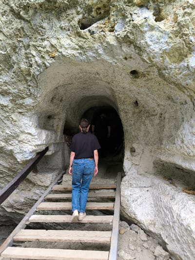 The image depicts the entrance to a man-made or natural cave system in Horaivka, Ukraine, carved into a light-colored rock formation. A single individual, viewed from behind, is ascending a set of wooden stairs leading into a dark, arched tunnel entrance. This person is wearing a black short-sleeved t-shirt, blue denim jeans, and light-colored closed-toe shoes. Their hair is dark and pulled back into a ponytail.
The stairs consist of rough-hewn wooden planks supported by a metal frame, with a dark metal handrail extending along the left side. The rock face surrounding the entrance is a textured, light grey to off-white material, possibly limestone or chalk, exhibiting irregular patterns and small, dark apertures in its upper sections. The interior of the tunnel beyond the immediate entrance is dark, with the faint outline of at least one additional figure discernible deeper within the gloom. The ground immediately around the base of the stairs consists of loose soil and rock debris. No direct interactions between individuals are visible. The environment appears to be a natural rock formation modified for access, indicative of a historical or natural site.