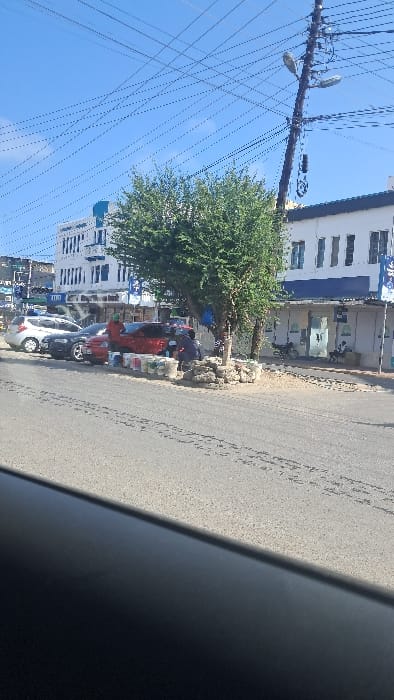 This image captures a daytime street scene in Majengo ward, Kenya, characterized by clear blue skies with numerous overhead power and communication lines crisscrossing the frame. On the right, a dark utility pole supports multiple wires and two streetlights.

The street, a paved asphalt road, shows tire marks and light debris. Vehicles are parked along the left curb; from left to right, these include a silver hatchback, a dark-colored sedan, and a red hatchback.

At the center-right of the image, two green-leafed trees grow on a raised dirt or paved area. Around the base of these trees, numerous white and colored buckets, consistent with paint containers, are arranged. Piles of light-colored rubble are also present near the tree bases.

Two individuals are actively engaged near the red hatchback and the paint buckets. One person, wearing a red top, stands beside the red car. Another person is seated or crouching near the tree, interacting with the buckets and rubble.

In the background, two multi-story white buildings with blue trim or branding line the street. The building on the left has "M-PESA" visible on its facade. The building on the right features a ground-level storefront with glass doors. A motorcycle is parked near this building's entrance, and a person is seated on a step or bench in front of it.