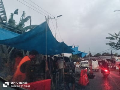 On a wet street in Kruet Teumpeun, Indonesia, construction or repair work appears to be underway under a blue tarp makeshift shelter. At least five workers are present, with one visible in an orange vest. Red safety barriers are deployed around the work area. A motorcycle is seen moving on the wet road. A white van and a wagon-like cart covered with a white tarp are parked in the background. A Pertamina gas station sign is visible in the distance. The sky is overcast, indicating rainy weather. Banana trees are visible on the left side of the image.