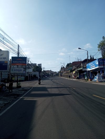 The image captures a straight, two-lane road in an urban setting. The road is asphalt, with yellow dividing lines. The left side of the road features a sidewalk with various businesses, including a Bank BJB ATM, a Kimia Farma pharmacy, and a Klinik Pratama (medical clinic). Utility poles with a dense network of wires are visible. On the right side of the road, shops with blue signage are present, including one that advertises "Jaringan XL" (an Indonesian telecom provider). A person is walking in front of this shop. A motorcycle is travelling in the center of the road. Further up the road, cars and trucks can be seen. The sky is a clear, bright blue. Shadows stretch across the road, suggesting a sunny day. The overall setting appears to be a main street in a bustling urban area.