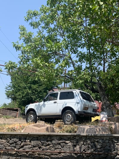 In the city of Karbi, Armenia, a white Lada Niva, an off-road vehicle, is parked on a dirt lot under a large, leafy tree. The car faces to the right, its rear end slightly angled towards the viewer. The vehicle appears to be in good condition, with its tires touching the dry, uneven ground. The area around the car is rough and unkempt, with patches of dry grass and a few scattered pieces of debris. In the background, a stone wall runs along the bottom of the frame, separating the dirt lot from what appears to be a lower level. Beyond the car, a dense green bush provides a backdrop, and a brown wooden structure is visible in the distance to the left. The sky is clear and blue, with the sun casting shadows.