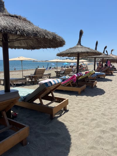 The image depicts a beach in San Roque, Spain, under a clear blue sky. In the foreground, several empty wooden lounge chairs with cushions are arranged on the sandy beach, some adorned with striped towels. Further back, more lounge chairs are occupied by people sunbathing or relaxing. Several thatched umbrellas provide shade over these chairs. The sea is visible in the background, with a few people wading in the shallow water and a distant pier with a lighthouse. The overall scene conveys a relaxed atmosphere of a sunny day at the beach.