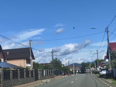 The image shows a street scene in Vișeu de Jos, Romania, under a partly cloudy blue sky. The road, an asphalt surface with white lane markings, curves slightly to the right. On the left side of the road, there is a stone and black metal fence in front of a house with a dark brown roof and a light pink facade. A modern-looking solar panel structure is visible over a portion of this property. On the right side of the road, there are several houses with red roofs, one with a white car parked in front. A large truck is driving away from the camera in the distance. Utility poles with numerous wires crisscross the sky above the street, and a streetlamp is visible on the right. Trees and vegetation are present along the sides of the road and behind the houses. There are a few people visible in the scene, including one on a red and white motorcycle on the left sidewalk, and a couple of figures near a truck on the right side of the road in the distance. Several road signs are also present.