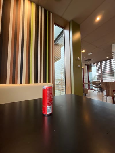The image captures the interior of a commercial establishment located in Wolfisheim, France. In the immediate foreground, a dark, reflective rectangular table is visible. Centrally positioned on the table stands an upright red 250ml Red Bull energy drink can, displaying a barcode and product information in multiple languages on its visible side. To the left of the table, a cream-colored upholstered booth backrest is partially visible. The left wall features a pattern of vertical stripes in varying widths and shades of brown, white, tan, and olive green. A large window, framed in wood, separates the striped wall from a light olive-green vertical pillar on the right. Through the window, an overcast exterior environment with bare tree branches and street furniture is discernible. Reflections of horizontal blinds are present on the window's surface. Beyond the pillar, an adjacent interior section is visible, containing additional seating arrangements including a curved wooden chair and a light-colored, round-backed chair on a pedestal base. The ceiling throughout the establishment is light-colored, featuring recessed lighting fixtures and a ventilation grate. No individuals are present in the scene.