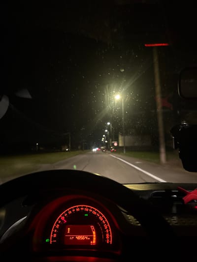 A nighttime car interior view features a red-lit dashboard with a speedometer displaying 48564 km. Raindrops dot the windshield, reflecting streetlights and car headlights ahead. A blurred steering wheel is visible. The road extends into the dark, wet night. The image captures a sense of driving in low visibility conditions.