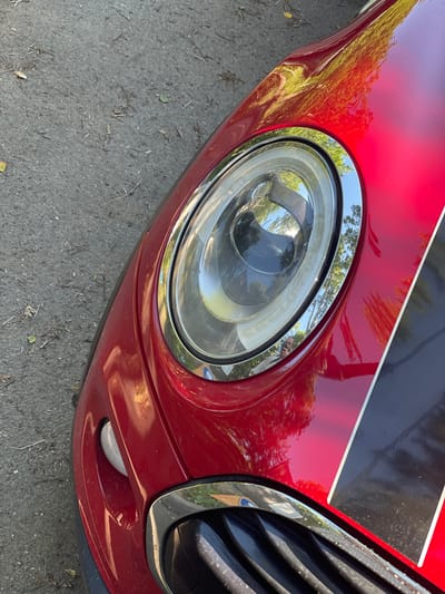 A close-up, high-angle shot focuses on the front quarter panel and headlight of a red car, likely a Mini Cooper, parked on a paved surface. The car's shiny red paint reflects the surrounding environment, including trees and sky, and the photographer is visible in a reflection on the headlight. A black graphic element with white trim is partially visible on the car's hood. The ground is a rough asphalt surface with scattered small debris, such as leaves and twigs. The scene is outdoors, with natural light illuminating the car and its surroundings. The location is identified as Sosnoviy Bor, Russia.