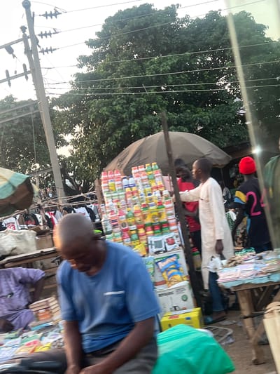 The scene depicts an active outdoor market in Toro, Nigeria, under a bright sky with visible utility poles and power lines above a canopy of green trees.
In the immediate foreground, a man with dark skin and a bald head sits, facing downwards and slightly to the right, wearing a blue short-sleeved t-shirt and dark trousers. To his left, another man in a purple traditional garment is seated, partially obscured, with stacks of books or papers in front of him. A green plastic bag rests on the ground near the seated man in blue.
In the mid-ground, a central market stall features a tall vertical display rack laden with numerous small, colorful bottles and containers, likely packaged food items or beverages. At the base of this display are cardboard boxes, including a yellow box and one marked "ROYALC," along with a package of "Always" brand chips. To the left of this display, an open umbrella with green and beige panels provides shade for a stall showcasing various t-shirts, one bearing the text "UNITED 33."
Several individuals populate the mid-ground. A man in a light-colored, long-sleeved tunic stands near the vertical merchandise display, gesturing with his left hand. Behind him, a man in a red t-shirt is partially visible, also near the display. To the far right, a man in a black t-shirt with large pink numbers "24" on the back and a red knitted hat stands. Further right, a wooden table covered with a light blue cloth displays multiple stacks of books, magazines, or papers. The ground beneath the stalls appears unpaved.