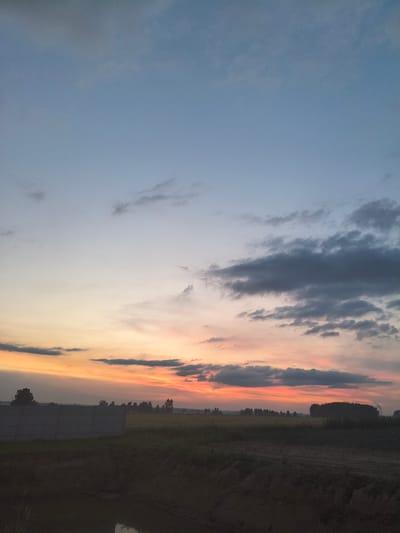 The image depicts a rural landscape at dusk. The sky transitions from a pale blue at the top to a gradient of soft orange, pink, and yellow near the horizon, illuminated by the setting sun. Scattered dark clouds drift across the sky. In the foreground, a dark, possibly muddy, area is visible, with some vegetation. A low, grey concrete wall runs horizontally across the middle ground, behind which a field or grassland stretches out. Silhouetted trees and a dense line of trees form the distant horizon. The overall impression is one of a peaceful evening in the countryside, possibly in or around Galonki, Poland.