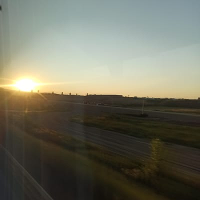 The image depicts a sunset over a rural landscape with a road and some structures in the distance. The sun is low on the horizon, casting a bright, warm glow across the sky and landscape, creating lens flares and a hazy effect. The terrain appears to be gently rolling hills, with areas of dry grass and dirt paths. In the middle ground, there is a road or track that curves through the landscape, and a few vehicles are visible. Further in the background, a line of buildings or structures is visible along the horizon, some appearing to be agricultural or industrial. The sky transitions from a pale yellow near the horizon to a light blue towards the top of the frame. The overall impression is one of a vast, open rural area at the end of the day.