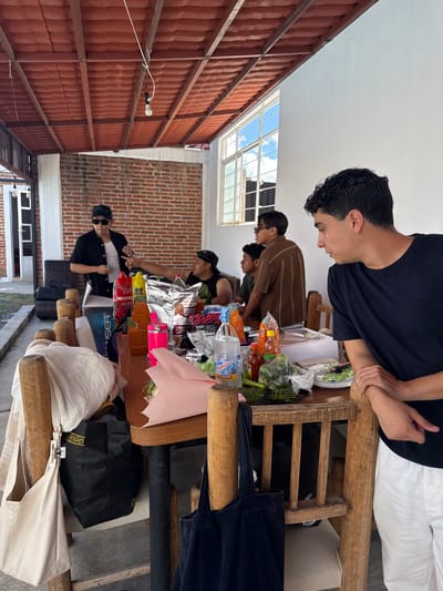 In San Baltazar Torija, Mexico, on an outdoor patio, several young men gather around a wooden table laden with food and drinks. The table is covered with various items including bags of snacks, bottles of soda, hot sauce, and what appears to be a bouquet of flowers. The men are casually dressed, some wearing hats and sunglasses. One man in a black shirt and white undershirt, wearing a black baseball cap and sunglasses, is standing and appears to be talking while holding a cigarette. Another man in a brown striped shirt is seated, looking towards the standing man. The patio is covered by a red tiled roof supported by wooden beams. A white wall forms the backdrop, and a window with blue skies and clouds is visible. The ground is a concrete pathway, and a garden area with grass is visible in the background.