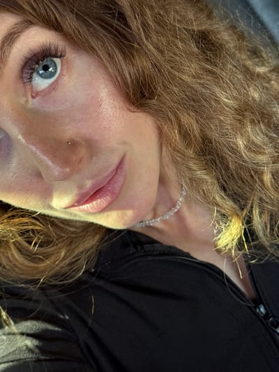 A close-up, angled shot focuses on a woman's face. Her eye, with defined lashes and a blue iris, is prominent. Her skin appears fair and smooth, with a visible nose piercing. Her lips are slightly parted, revealing a hint of pink. Her hair is thick, curly, and a mix of light brown and blonde shades. A delicate necklace rests on her neck. She is wearing a black garment with a zipper visible near her neck. The lighting is a mix of bright and shadowed areas, giving the image a slightly dramatic effect. There is no evidence to suggest that the photo was taken in Machara, Abkhazia.
