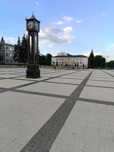 The image depicts a wide, paved public square in Gubkin, Russia, dominated by a tall, ornate clock tower in the foreground. The square is laid with light-colored paving stones arranged in a grid pattern, accented by darker, textured strips. The clock tower, featuring four faces and a decorative spire, stands prominently to the left. In the background, a modern, light-colored building with multiple windows and a flat roof is visible, surrounded by trees. Several individuals are scattered across the square, some walking, others appearing to be resting or engaged in activities like riding scooters. The sky above is blue with scattered white clouds, suggesting daytime. The overall scene conveys a sense of an urban public space during a clear day.