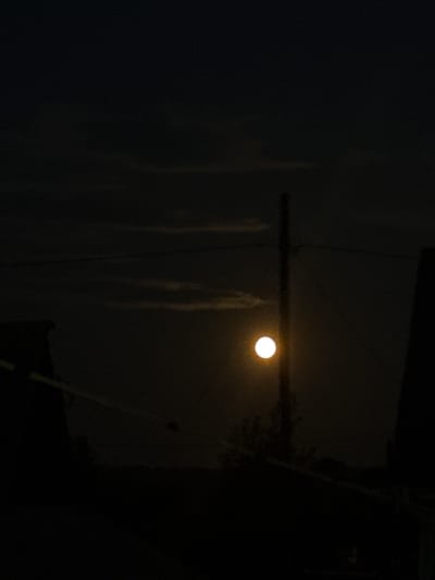 A vertical, full shot captures a dark, nighttime scene with a bright, full moon as the primary light source. The moon, a distinct, glowing orb, is positioned slightly left of center, casting a warm, yellowish light that illuminates the surrounding clouds with a faint glow. In the foreground, silhouetted structures, possibly rooftops or buildings, are visible in deep shadow, with the dark outlines contrasting against the lighter sky. A tall, thin pole or post stands vertically near the center, partially obscuring the moon and creating a strong vertical line in the composition. Faint horizontal lines, likely power or telephone wires, stretch across the frame, adding a sense of depth and urban or semi-urban environment. The overall atmosphere is somber and quiet, dominated by the darkness of night and the ethereal glow of the moon. There is no indication of the specific location within Baryshevo, Russia, beyond it being a nighttime urban or semi-urban setting.