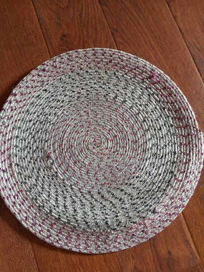 A round, woven placemat with a spiral pattern is placed on a wooden floor. The placemat is predominantly silver with accents of black, red, and purple threads. The wooden floor consists of planks with a reddish-brown hue and a visible grain. The image appears to be taken indoors, with the lighting suggesting an overhead source. There are no people or animals present in the image. The location in the city of Vilyuysk, Russia, is not discernible from the visual information provided in the image.