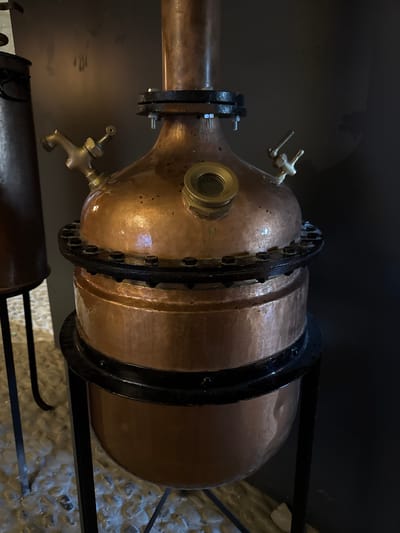 The image shows a close-up, slightly elevated, eye-level view of a copper alembic, a piece of distillation equipment, likely used for producing spirits or essential oils. The alembic is made of polished copper and features a bulbous lower section connected to a cylindrical upper section via a flanged joint secured with bolts. Two brass spigots or valves are visible on the upper bulbous part, one on the left and one on the right. A round sight glass with a brass housing is also present on the upper bulbous section, positioned centrally. The alembic stands on a black metal frame. To the left, a portion of another dark metal cylindrical object is visible, also on a dark metal stand, with a handle. The background is a dark, nondescript wall, and the floor appears to be covered in light-colored, uneven stones or pebbles. The lighting is dim, highlighting the metallic sheen of the copper. The scene is indoors, suggesting a workshop, distillery, or a display in a historical or museum setting. The location is identified as Dolceacqua, Italy.