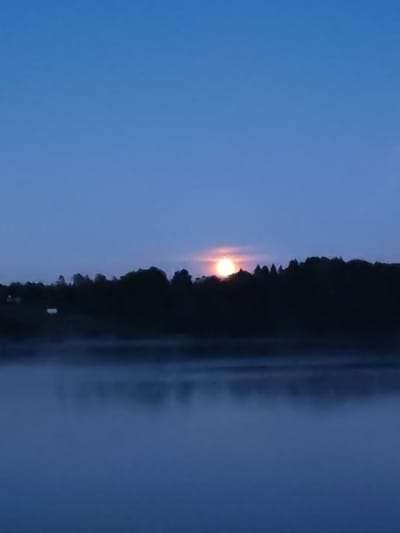 The image depicts a low-light landscape scene located in Irma, Russia. The upper third of the frame shows a clear, deep blue sky. A dark, undulating silhouette of a forested landmass or tree line spans the middle ground, extending horizontally across the image. Centered above this silhouette, a bright, circular, orange-yellow orb, consistent with the moon or sun near the horizon, is partially visible, emitting a distinct glow. The lower two-thirds of the image portray a body of water, likely a lake or river, partially obscured by a layer of low-lying mist or fog that covers its surface. Faint, dark reflections of the landmass and a diffuse glow from the orb are discernible on the water's surface beneath the mist. No individuals, animals, specific actions, or interactions are present within the scene. The environment appears natural and undisturbed.