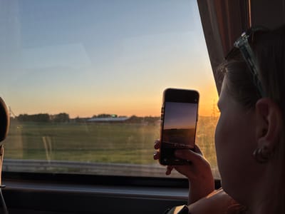 A woman sits in a vehicle, possibly a bus or train, looking out of a window at dusk. She holds a smartphone, using it to take a picture of the view, which includes a field and buildings in the distance. The sky is a gradient of blue and orange, indicating the setting sun. The location is likely Bol'shiye Klyuchishchi, Russia, based on associated information.