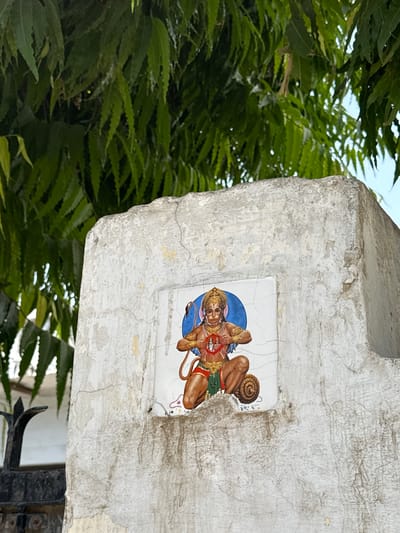 The image depicts a weathered, rectangular concrete structure, possibly a pillar or support, on which a ceramic tile depicting a deity is affixed. The tile is rectangular and features a depiction of Hanuman. The background of the tile is a blue circle and the cracks in the tile suggest its age and exposure to the elements.
The concrete structure itself shows signs of age and wear, with cracks and uneven surfaces. It is situated outdoors, as evidenced by the lush green foliage visible in the background. The leaves suggest the presence of trees or bushes, contributing to the outdoor environment.
The photograph was taken in Mathura, India.