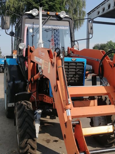 The image shows a close-up, ground-level, eye-level view of a blue and orange tractor with a front-end loader, parked outdoors. The tractor is primarily blue on its rear section and the front loader is a bright orange. The tractor has a large, thick tire visible on the left side. The cab of the tractor is partially visible, with windows and mirrors. On the orange loader, there is visible text in Russian, including "БЛЮМИНГ" and "БД-750". The background consists of trees, a blue sky, and a portion of an industrial-looking building structure. The ground appears to be paved or a hard surface, with some dark spots that may indicate oil stains. The scene is consistent with a tractor parked at a service area or an agricultural facility. The text on the loader suggests the tractor is a product or has attachments from a company in Belarus. The location is identified as Zhabinka, Belarus, a city known for its agricultural and industrial activities. There are no people or animals visible in the image. The lighting suggests it is daytime, with sunlight casting shadows.