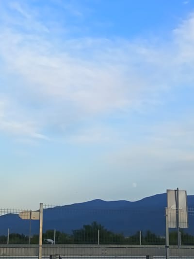 The image depicts a serene natural landscape with a clear blue sky and soft white clouds. In the distance, a mountain range forms a dark blue silhouette against the horizon. A pale orb, possibly the moon, is visible in the sky above the mountains. In the foreground, a chain-link fence runs horizontally across the frame, separating the viewer from the landscape. A white sign on a pole stands to the right of the fence. The setting appears to be a quiet, outdoor location.