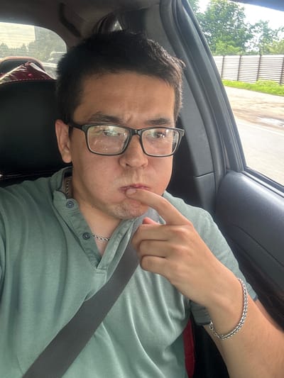 The image shows a person in the passenger seat of a car. The person is wearing glasses, a light green collared shirt, and a silver chain necklace. They are also wearing a silver bracelet on their right wrist. The person's right index finger is touching their chin, and they are looking towards the camera with a neutral expression. The car's interior is visible, including a black seat and a seatbelt. Through the car window, trees and a fence can be seen, suggesting an outdoor setting. The lighting indicates it is daytime. The context provided places the location as Kuzmolovskiy, Russia.