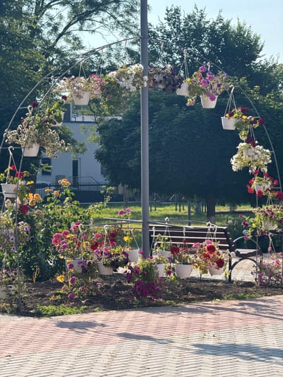 The image shows a large floral archway adorned with numerous hanging flower baskets in various colors, including pink, red, yellow, and white. The archway is made of metal and is supported by a central pole. In the background, there is a building, trees, and a park bench. The ground in the foreground is paved with brick. The scene appears to be a public park or garden in the city of Ungheni, Moldova. There are no people visible in the image.