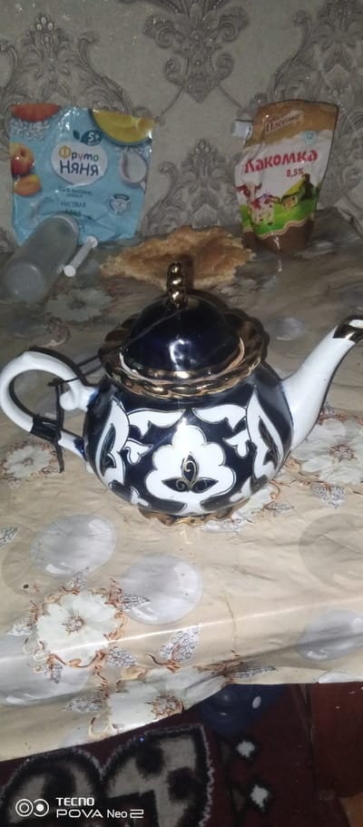 The image depicts a still life arrangement on a patterned tablecloth. In the foreground, a blue and white patterned teapot with gold accents is the central subject. To the left of the teapot, a clear plastic bottle lies on its side. Behind the teapot, a package of baby food and a pouch of what appears to be condensed milk are visible. A piece of flatbread rests between the food items. The background features a textured wallpaper with a damask pattern. The scene is illuminated by artificial light, casting subtle shadows. There is no indication of location within the city of Tahmach, Uzbekistan.