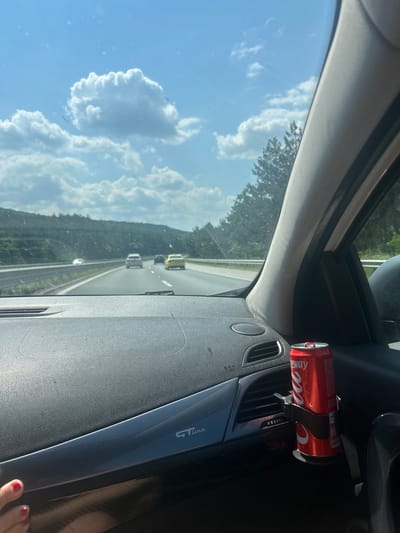 This image was taken from the interior of a car traveling on a highway. The sky is blue with white clouds. The highway has multiple lanes and is surrounded by trees and greenery. There are several cars on the highway. In the foreground, on the dashboard of the car, is a red can of Coca-Cola. A portion of a person's hand with red nail polish is visible on the left side of the image. The location of Novi han, Bulgaria is mentioned, but there is no specific visual information in the image that confirms this location.