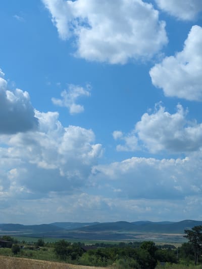 The image captures a scenic view of the Bulgarian countryside, likely near the village of Sokolovo. The foreground is dominated by a field of dry, golden grass, possibly harvested wheat or barley, stretching across the bottom of the frame. Beyond this field, a band of lush green vegetation marks the transition to rolling hills and distant mountains. The landscape is dotted with scattered trees and a few small buildings, suggesting a rural setting. The sky above is a vibrant blue, filled with a variety of clouds, from fluffy white cumulus formations to more scattered, wispy clouds, creating a dynamic and expansive atmosphere. The overall impression is one of a peaceful, natural environment under a bright, partly cloudy sky.