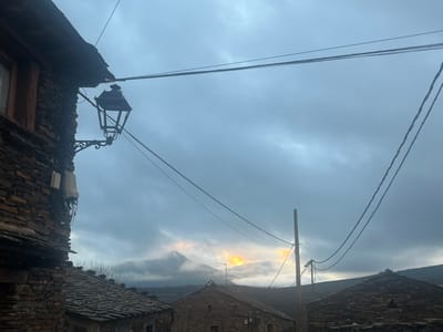 The image captures a rural village scene in Campillo De Ranas, Spain, characterized by traditional dark stone architecture under an overcast sky. In the foreground, on the left, a building constructed from dark, irregular stone blocks features a window with a wooden frame. A decorative black metal street lamp, suspended by an ornate bracket, protrudes from this building's wall, with various utility boxes and wires clustered beneath it.

Numerous power lines traverse the frame, stretching across the sky and converging towards a wooden utility pole positioned on the right side of the image. Below these lines, the distinctive dark slate roofs of other village houses are visible, blending with the somber tones of the landscape.

In the mid-ground and background, a misty mountain peak rises, partially veiled by clouds. Behind this mountain, a striking golden light, likely from the rising or setting sun, breaks through the heavy grey cloud cover, casting a warm glow on the horizon and creating a dramatic contrast with the darker foreground. The overall impression is one of a quiet, possibly cool, early morning or late evening in a mountainous, rustic setting. No people or visible text are present.
